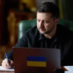 Володимир Зеленський сидить за столом і пише в блокноті біля ноутбука з прапором України