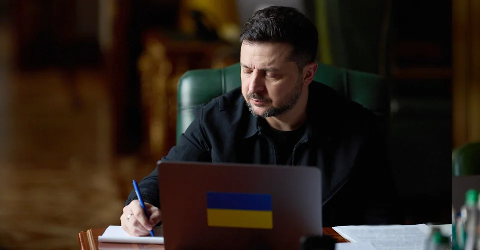 Володимир Зеленський сидить за столом і пише в блокноті біля ноутбука з прапором України