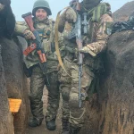 Два українські військові в камуфляжі стоять в окопі зі зброєю