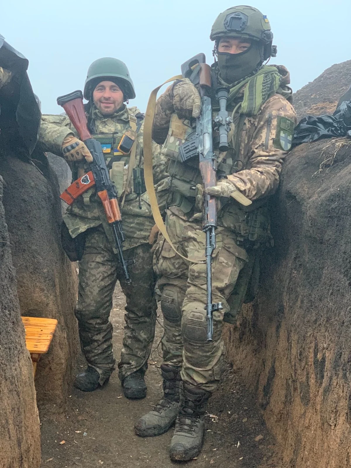 Два українські військові в камуфляжі стоять в окопі зі зброєю