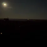Нічне небо з яскравим місяцем над темним силуетом місцевості з поодинокими вогнями