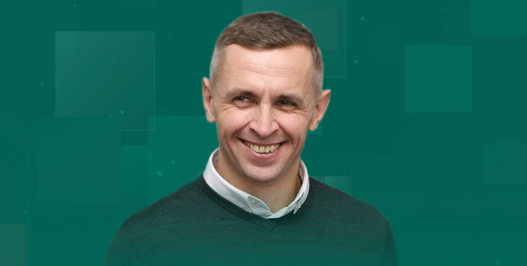 Усміхнений чоловік з коротким волоссям у зеленому светрі поверх білої сорочки на зеленому тлі