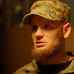 Чоловік у камуфляжній формі та кепці з рудою бородою дивиться вбік